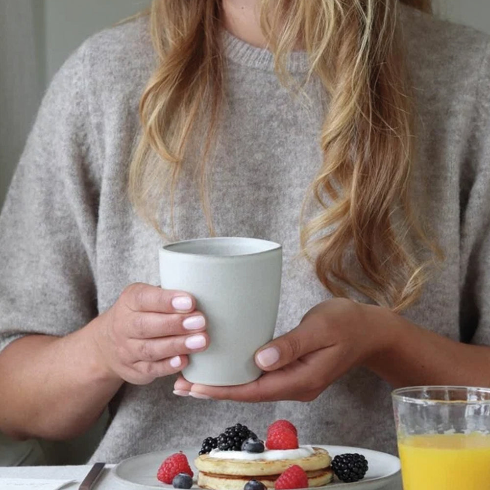 vrouw met een grijze mok in haar hand en ontbijtproducten op tafel