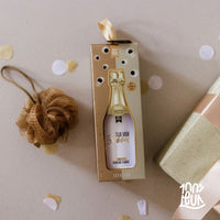 Een showerpuf en een geschenkset met een douchegel erin in een gouden doosje. Er liggen wat confetti's bij op een beige achtergrond. 