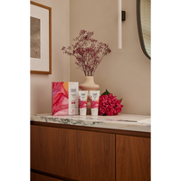 Therme Blooming Selfcare geschenkset met douchegel en bodylotion op een marmeren badkamermeubel, naast roze bloemen en een vaas met droogbloemen. Een sfeervol verzorgingscadeau voor een verwenmoment thuis.