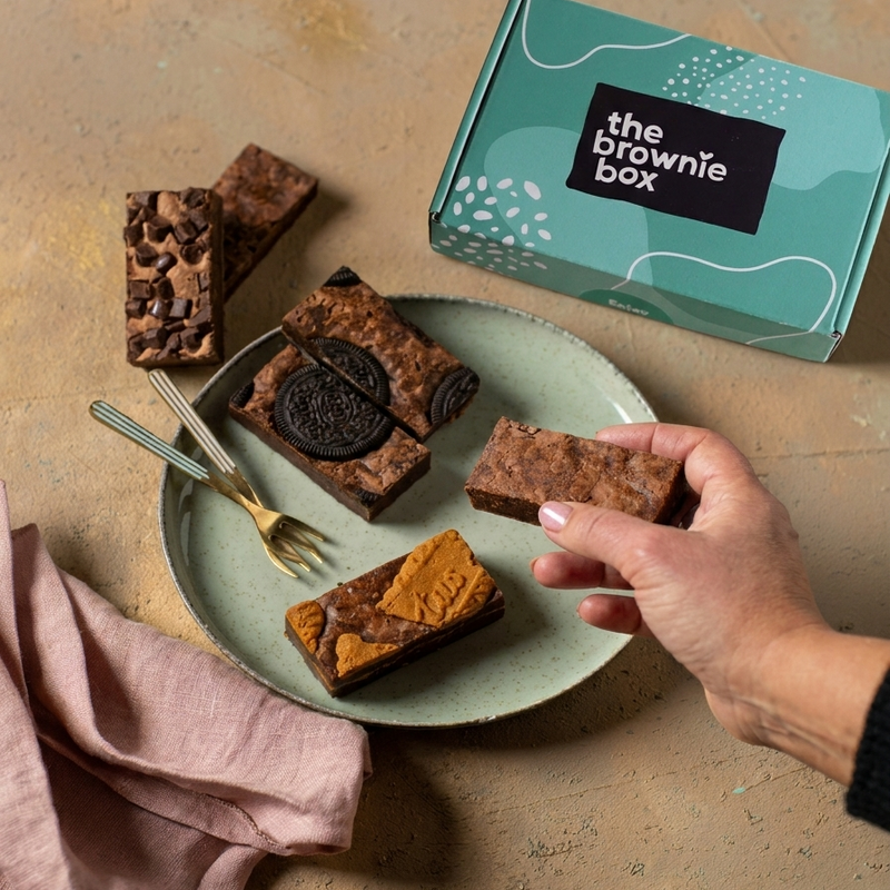 vegan oreo brownies, vegan originals brownies, vegan speculoos brownies en vegan double chocolates brownies op een bordje met The Brownie Box doosje erbij op een rustige achtergrond